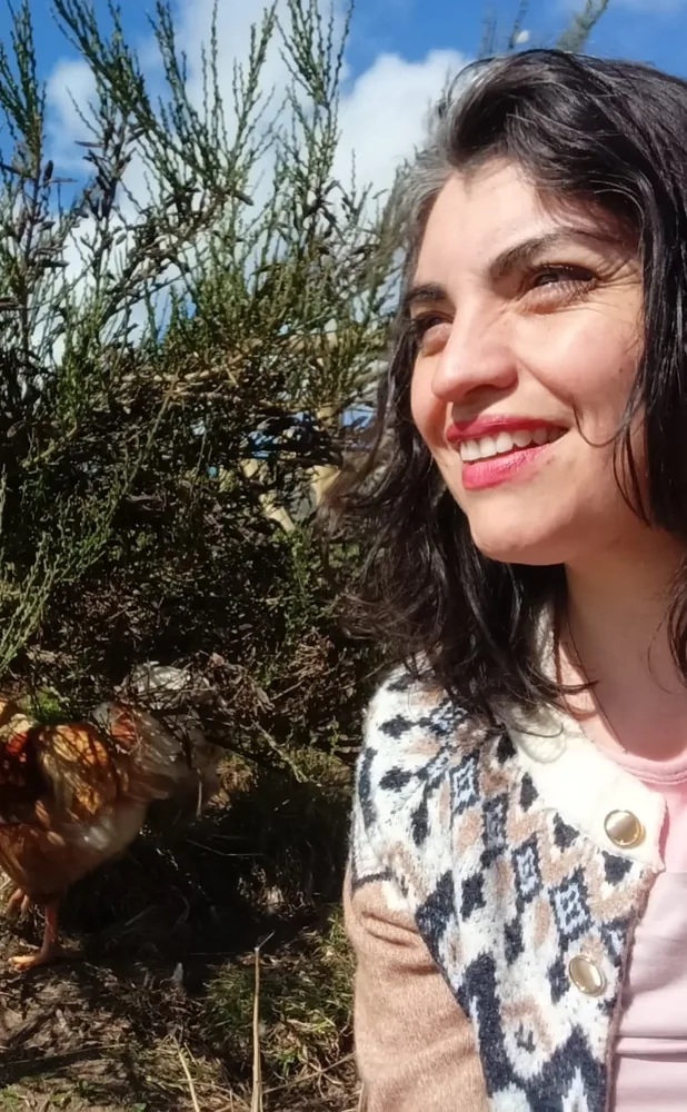 Maritza sonríe al sol junto a un arbusto, con una gallina picoteando a su lado