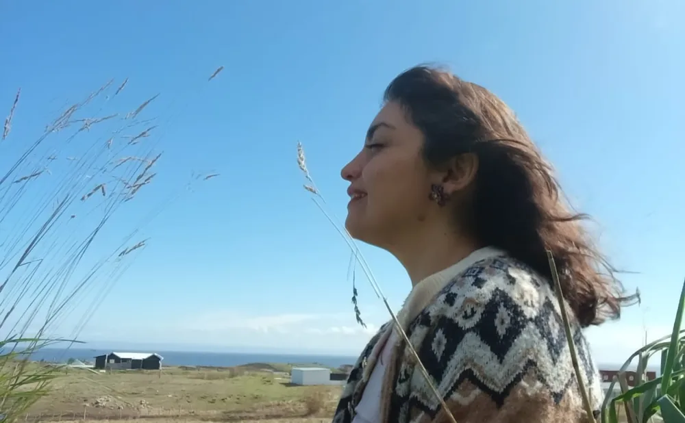 Maritza de perfil, con los ojos cerrados y el pelo al viento, entre pastos altos frente a la estepa y el mar